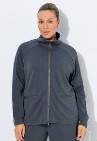 Mujer vestida con chaqueta deportiva gris oscuro con cremallera y pantalones a juego, de pie contra una pared blanca, con el cabello recogido.