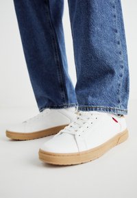 Białe sneakersy z fakturowanej skóry, gumową podeszwą, z logo po boku, połączone z jasnoniebieskimi dżinsami.