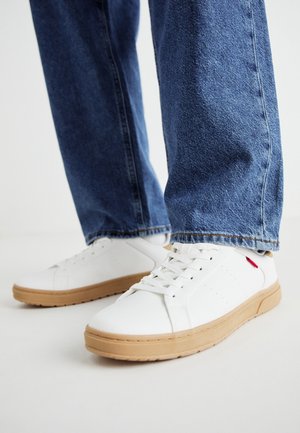Białe sneakersy z fakturowanej skóry, gumową podeszwą, z logo po boku, połączone z jasnoniebieskimi dżinsami.
