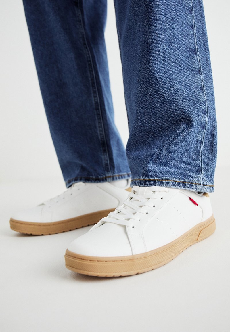 Białe sneakersy z fakturowanej skóry, gumową podeszwą, z logo po boku, połączone z jasnoniebieskimi dżinsami.