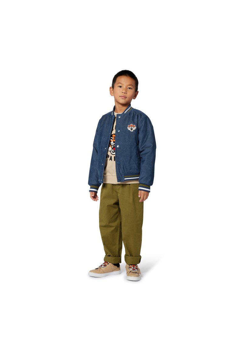 Veste bomber en denim avec des poignets et un col côtelés, arborant un patch brodé. T-shirt graphique beige, pantalon vert olive et baskets beiges avec des accents.