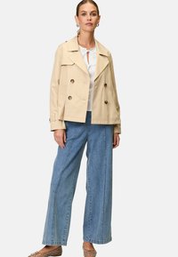 Beige dubbelknäppt jacka med stora knappar, i sällskap med en vit blus och vida blå denimbyxor. Stilen är avslappnad.