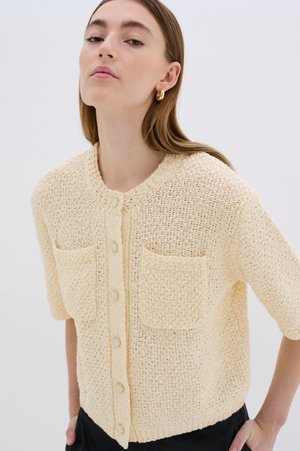 Light beige knitted cardigan with short sleeves, two front pockets, and round neckline. Features large buttons and a textured fabric.