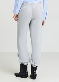 Persona che indossa pantaloni sportivi grigi chiari con polsini elastici alle caviglie, una maglietta a maniche lunghe blu chiaro e stivaletti neri, in piedi su un pavimento semplice.