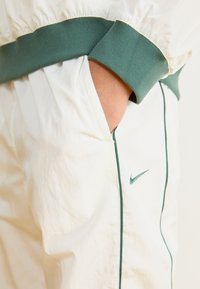 Baltie sporta bikses ar zaļiem akcentiem, ar mīkstu elastīgu jostasvīru un aprocēm, Nike logo izšūts uz augšstilba, redzama sānu kabata.