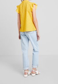 Yellow short-sleeve top with a floral eyelet design, paired with light blue rolled-up trousers and white block-heeled sandals.