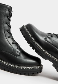 Las botas de cuero negro hasta el tobillo presentan una puntera redondeada, cierre de cordones y una suela gruesa de goma con una textura de dibujo y costuras decorativas.