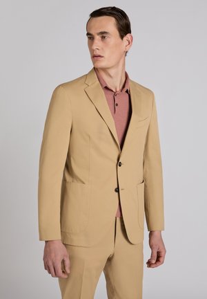 Homme portant une veste de costume beige et un pantalon assorti avec un polo rouge pâle, debout devant un fond gris clair uni, regardant vers la gauche.