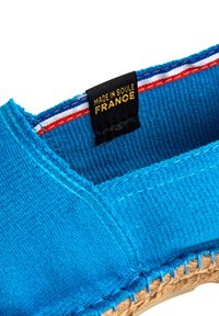 Espadrille en toile bleue avec une semelle en jute tissée. Présente une étiquette noire indiquant "FABRIQUÉ EN SOLE FRANCE" avec des accents rayés rouge et blanc.