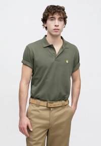 Πόλο πουκάμισο σε ελιά πράσινο με γιακά, γυρισμένα μανίκια και μικρό κίτρινο λογότυπο. Συνδυάζεται με μπεζ παντελόνι και ζώνη στο ίδιο χρώμα.