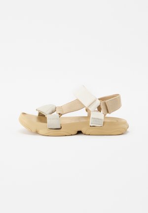 Sandali sportivi beige con suola in gomma flessibile, cinturini in tessuto regolabili e supporto imbottito per la caviglia; design minimalista, forma piatta e tomaia testurizzata.