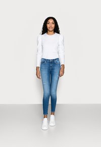 Witte lange mouwen top met een kantenkraag, gecombineerd met lichtblauwe skinny jeans en witte sneakers, op een neutrale achtergrond.