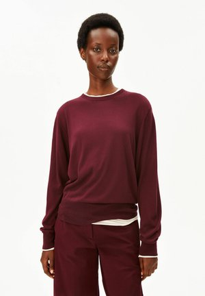 Femme aux cheveux courts naturels portant un pull rouge foncé à manches longues et un pantalon assorti, debout devant un fond blanc uni.