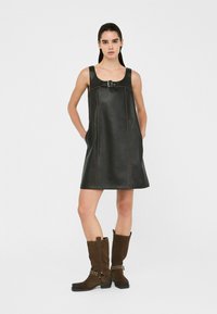 Abito A-line in pelle nera con spalline larghe, caratterizzato da un dettaglio a fibbia e cuciture marroni a contrasto. Abbinato a stivali al ginocchio in suede marroni.