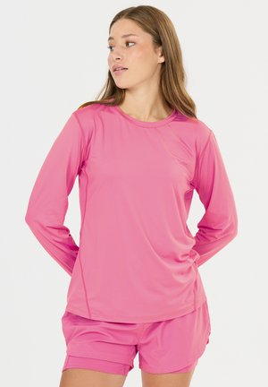 Frau trägt ein knallpinkes Langarmshirt und passende Shorts, schaut nach links vor einem schlichten weißen Hintergrund.