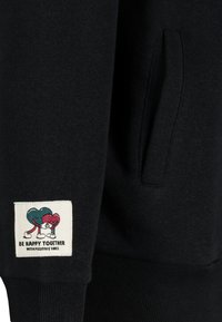 Zwarte sweatshirt met een zachte textuur, voorzien van een zak en een label met een kleurrijke illustratie van twee hartpersonages die elkaar omarmen.