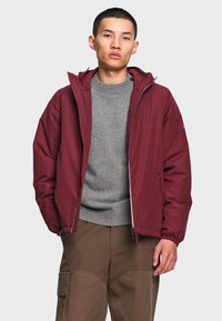 Jeune homme portant une veste bordeaux zippée sur un pull gris et un pantalon marron, debout avec une main dans la poche devant un fond uni.