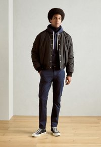Tume bomber-jakk hõbedaste nööpidega, sinine kapuutsihoodie all, sinised cargo-püksid ja mustad-valged tossud puidust põrandal.