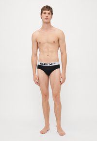 Junger Mann, barfuß, trägt schwarze Unterhosen mit weißem Bund und kräftigem schwarzen Text in einem hellen, minimalistischen Studio.