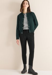 Tmavě zelená bomber bunda, šedý svetr pod ní, černé strukturované kalhoty a černé šněrovací boty. Hladká látka a přiléhavý střih.