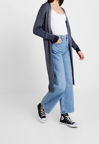 Lång marinblå kofta med ribbad struktur, öppen front och kontrasterande mörka manschetter, bärs över en vit tanktop och ljusblå vida byxor med svarta sneakers.