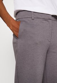 Grå byxor i en strukturerad tyg, med rak passform, sidofickor och en platt midja, med synlig handplacering.