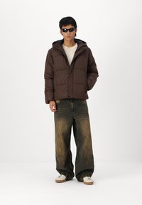 Brun polstret jakke med hætte, iført over en beige skjorte. Kombineret med løse, faded blå jeans og hvide sneakers med brune såler.