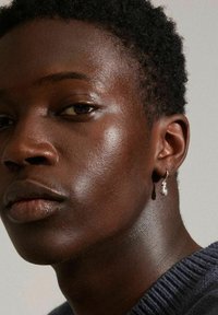 Boucle d'oreille argentée avec un détail en perle en forme de goutte d'eau, portée par une personne à la peau lisse et sombre, avec des cheveux courts et texturés, portant un pull sombre.