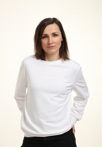 Frau mit schulterlangem braunem Haar, die einen weißen Sweatshirt und schwarze Hose trägt, steht mit den Händen hinter dem Rücken vor einem schlichten Hintergrund.