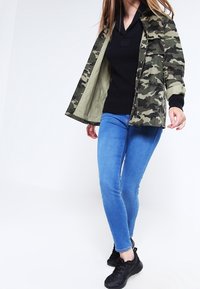 Jacket s maskáčovým vzorem v zelené a béžové barvě, nošený přes černý pletený svetr, spárovaný s modrými skinny džínami a černými sportovními botami.