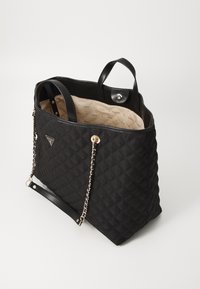 Sac cabas matelassé noir avec des anses chaîne dorées, double poignées, et intérieur beige imprimé de logos "GUESS", ouverture supérieure avec fermeture à clip.