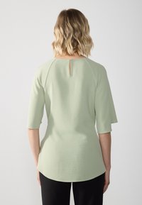 Femme aux cheveux blonds ondulés mi-longs portant un haut texturé vert clair avec des manches aux coudes et une fermeture dos en forme de goutte d'eau.