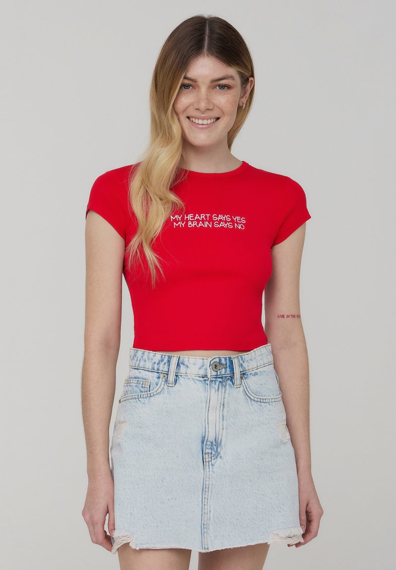 T-shirt rosso aderente con maniche corte, caratterizzato da testo bianco sul petto, abbinato a una mini gonna di denim blu chiaro con orlo sfrangiato.