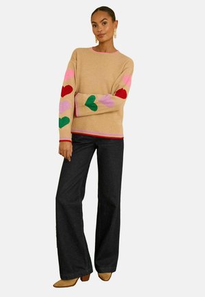 Knit sweater in beige with colorful heart patterns in pink, red, and green on sleeves, and pink accents. Paired with dark denim pants and tan heels.