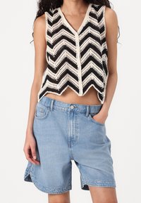 Černobílá pletená vesta se vzorem chevron a výstřihem do V, doplněná světle modrými džínovými šortkami s bočními kapsami a volným střihem.