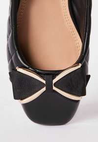 Zapato plano negro con parte superior de cuero sintético acolchado, plantilla nude y acento de lazo negro con un detalle metálico dorado en la punta.