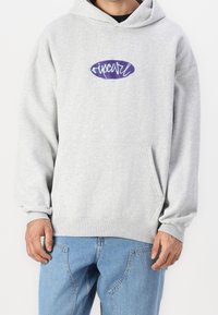 Szara bluza z kapturem, dużą przednią kieszenią i luźnym fasonem. Na piersi fioletowe logo graficzne. Materiał z mieszanki bawełny.