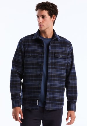 Chemise en flanelle à carreaux bleu et noir avec un devant boutonné, deux poches poitrine et un ourlet arrondi. Texture douce, manches longues et col.