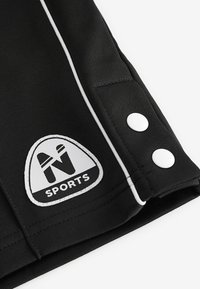 Sorte sportsbukser med hvide detaljer, der har et trekantet logo-patch og to hvide trykknapper i sidesømmen. Glat tekstur.