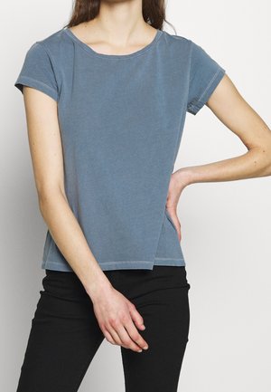 Femme portant un t-shirt bleu délavé à manches courtes et un pantalon noir, posant avec une main sur la hanche et l'autre reposant devant.