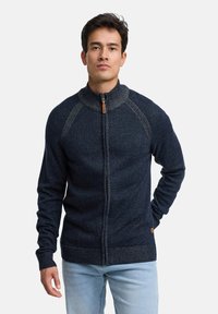 Granatowy sweter zapinany na zamek z wysokim kołnierzem, dzianą teksturą i rękawami typu raglan. Posiada mały skórzany suwak oraz ściągacze na mankietach.