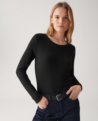Junge Frau mit blonden Haaren, die ein schwarzes, geripptes Langarmoberteil und dunkle Jeans mit einem schwarzen Gürtel trägt, mit den Händen in den Taschen, neutraler Hintergrund.