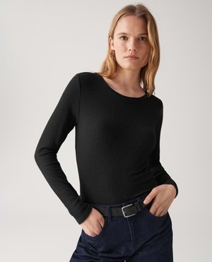 Giovane donna con capelli biondi che indossa una maglietta a maniche lunghe nera a costine e jeans blu scuro con una cintura nera, mani nelle tasche, sfondo neutro.