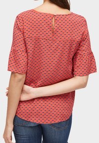 Punainen toppi, jossa sininen geometrinen kuvio, lyhyet liehuvat hihat, avonaistakakappale ja kaareva helma. Yhdistetty sinisiin farkkuihin.