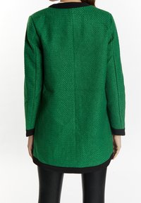 Abrigo verde texturizado con un corte elegante, detalles negros en el cuello y el dobladillo, con mangas largas y un ajuste entallado.