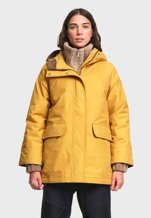 Femme portant une veste d'hiver jaune à capuche par-dessus un pull beige zippé, debout face à l'avant, les bras détendus le long du corps.