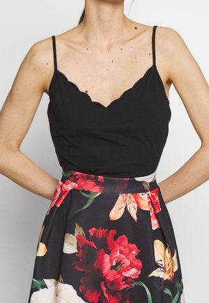 Kvinde iført en sort camisole med bølget kant kombineret med et blomsterprintet skørt med store røde og orange blomster.