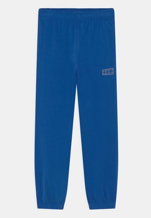 Pantalon de survêtement bleu à taille élastique avec chevilles resserrées et petit logo blanc en forme de grille rectangulaire sur la partie supérieure de la cuisse gauche.