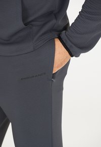 Graue Sporthose aus strukturiertem Stoff mit Logodruck, seitlicher Tasche und tailliertem Design. Die Ecken weisen gerippte Details auf.
