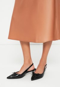 Zwarte leren slingback schoenen met spitse neuzen en decoratieve strikken, gecombineerd met een gladde, midi-lange perzikrok.